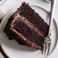 A slice of moist layered Devil's Food Cake with rich chocolate buttercream frosting on a white plate.
